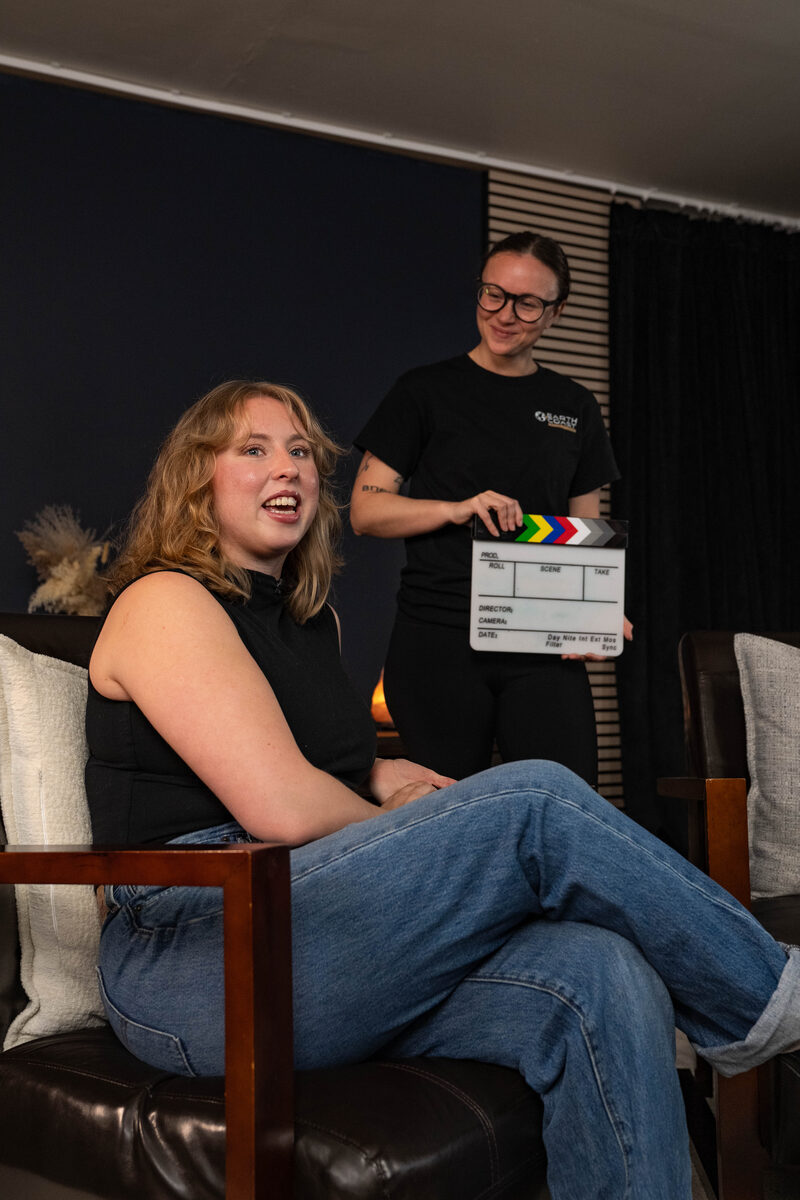 A guest laughs on a leather armchair in the Earth Coast Studios lounge while a crew member in an Earth Coast tee holds a clapperboard.